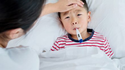 Bé nằm trên giường với nhiệt kế trong miệng trong khi mẹ đặt tay lên trán