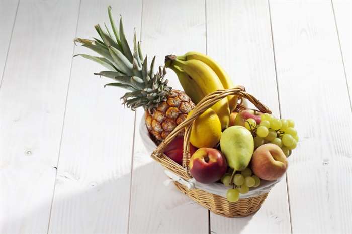 Giỏ trái cây với dứa, chuối, chanh, táo, đào, nho trên nền gỗ trắng.