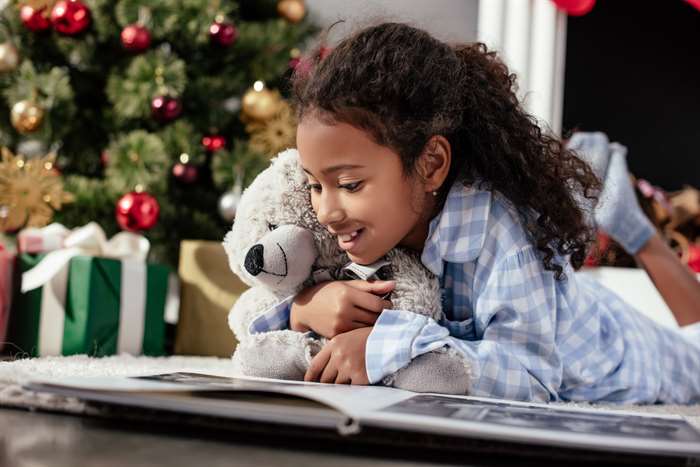 happy adorable child in pajamas with teddy bear looking at photo album on floor at home