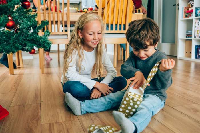 Two kids sitting on the floor and opening christmas gifts near the Christmas tree.