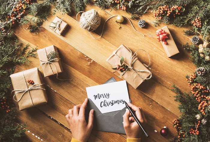 Người viết thiệp Giáng sinh trên bàn gỗ bên cạnh những món quà được gói và vòng hoa.
