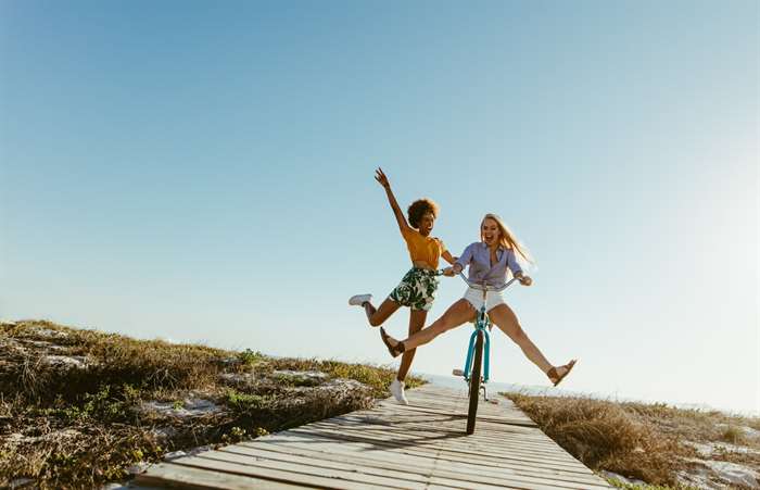 Excited woman riding bike down the boardwalk with her friends running behind. Two young female friends having a great time on their vacation.